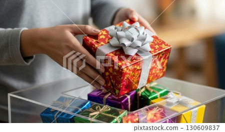 Person holding decorated gift box with bow in festive setting, colorful presents in background 130609837