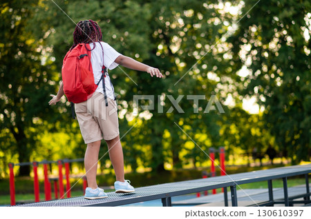 Girl with a red backpack walking on the log on a playground and keeping balance 130610397