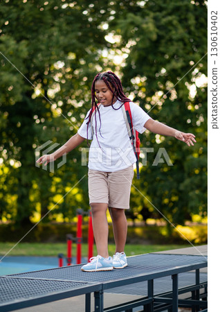Girl with a red backpack walking on the log on a playground and keeping balance 130610402