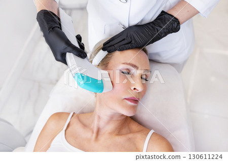 Cosmetologist in black gloves performing hardware facial treatment with modern device on mature woman lying in clinic. Concept of beauty, anti-aging, dermatology, skincare, rejuvenation Cosmetologist in black gloves performing hardware facial treatment with modern device on mature woman lying in clinic. Concept of beauty, anti-aging, dermatology, skincare, rejuvenation 130611224