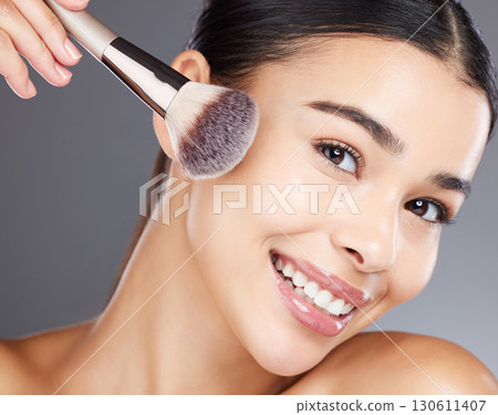 Face, beauty makeup and woman with brush in studio isolated on a gray background. Portrait, aesthetics and happy young female model apply powder, foundation or cosmetics product for skincare wellness 130611407