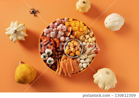 Halloween sweet charcuterie board with pumpkin, popcorn, dried fruits, color chocolate candy, spider, crackers in white plate on orange background. Festive food for kids party. Top view. Halloween sweet charcuterie board with pumpkin, popcorn, dried fruits, color chocolate candy, spider, crackers in white plate on orange background. Festive food for kids party. Top view. 130612309