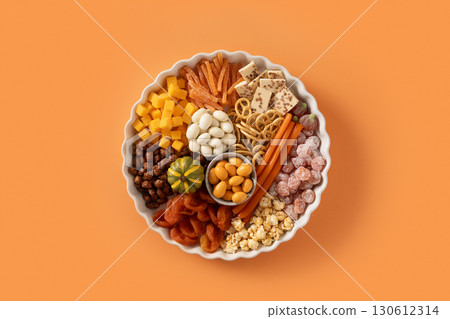 Halloween sweet charcuterie board with pumpkin, popcorn, dried fruits, color chocolate candy, spider, crackers in white plate on orange background. Festive food for kids party. Top view. Halloween sweet charcuterie board with pumpkin, popcorn, dried fruits, color chocolate candy, spider, crackers in white plate on orange background. Festive food for kids party. Top view. 130612314