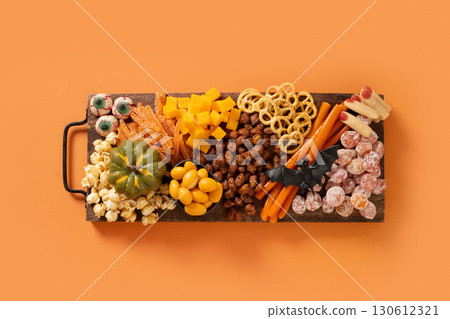 Halloween sweet charcuterie board with pumpkin, popcorn, dried fruits, color chocolate candy, spider, crackers on cutting board on orange background. Festive food for kids party. View from above. Halloween sweet charcuterie board with pumpkin, popcorn, dried fruits, color chocolate candy, spider, crackers on cutting board on orange background. Festive food for kids party. View from above. 130612321