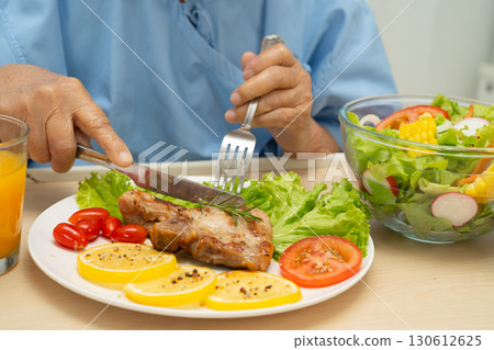 Asian senior woman patient eating pork chop stake and vegetable salad for healthy food in hospital. 130612625