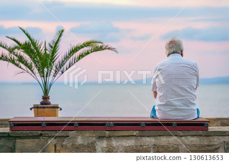 Elderly man sitting alone on bench at seaside promenade, facing ocean under evening sky. Concept of solitude, senior lifestyle, peaceful moment of reflection 130613653
