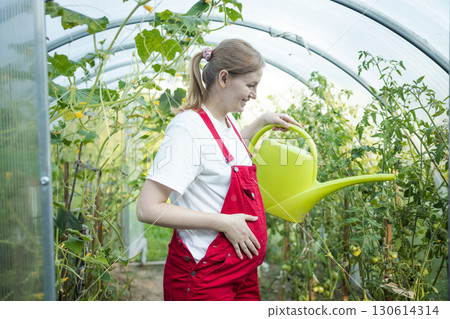 Pregnant woman watering plants in greenhouse with watering can 130614314