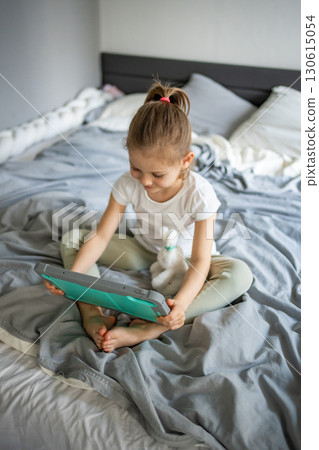 Little girl using a tablet while sitting on her parents bed in the bedroom. Concept of screen time, digital play and modern childhood in a home environment. 130615054
