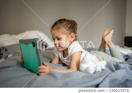Little girl lying on her parents bed and watching cartoons on a tablet. Concept of screen time, digital entertainment and relaxation during early childhood. Little girl lying on her parents bed and watching cartoons on a tablet. Concept of screen time, digital entertainment and relaxation during early childhood. 130615078