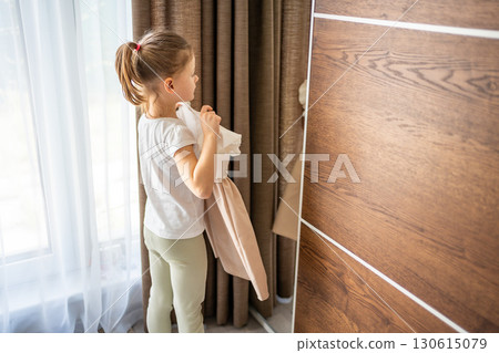 Little girl standing in front of a mirror and holding a dress against herself. Concept of self-expression, imagination and early interest in clothing and appearance. Little girl standing in front of a mirror and holding a dress against herself. Concept of self-expression, imagination and early interest in clothing and appearance. 130615079