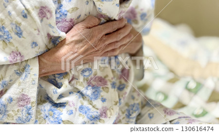 An elderly woman wearing pajamas and holding her chest with both hands 130616474