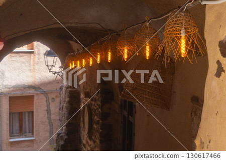 Warm lanterns with woven shades light up a rustic stone alleyway, creating a cozy and inviting ambiance at dusk. The charming old-world architecture enhances the scene's enchanting mood Warm lanterns with woven shades light up a rustic stone alleyway, creating a cozy and inviting ambiance at dusk. The charming old-world architecture enhances the scene's enchanting mood 130617466