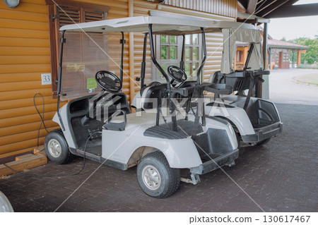 Two white golf carts are parked under a wooden shed next to a log cabin. The carts are plugged in for charging, indicating preparation for future use or maintenance. Two white golf carts are parked under a wooden shed next to a log cabin. The carts are plugged in for charging, indicating preparation for future use or maintenance. 130617467