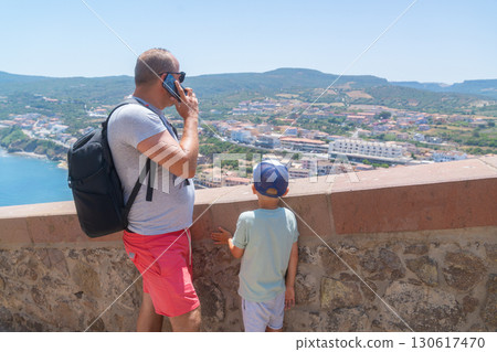 Father and son stand at a viewpoint overlooking a coastal town. The father talks on a phone while the son looks at the landscape, showcasing a blend of modern technology and family connection. Father and son stand at a viewpoint overlooking a coastal town. The father talks on a phone while the son looks at the landscape, showcasing a blend of modern technology and family connection. 130617470