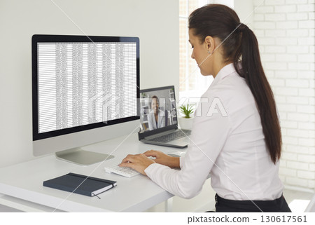 Accountant working with spreadsheets on desktop computer during video call with client on laptop 130617561