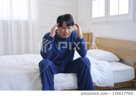 A man in his 20s sitting on the bed with a headache 130617760
