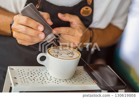 Barista decorating cappuccino with chocolate syrup, creating detailed latte art on foamy surface. Concept of coffee art, barista skills, creative drink preparation 130619808