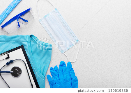 Health care background of disposable face mask gloves cap goggles scrubs and stethoscope and clipboard laying on table flat lay view 130619886
