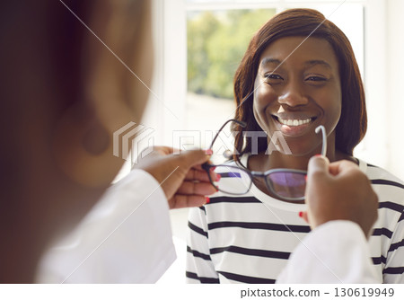 Professional optician offers new eyeglasses to happy smiling young African woman Professional optician offers new eyeglasses to happy smiling young African woman 130619949