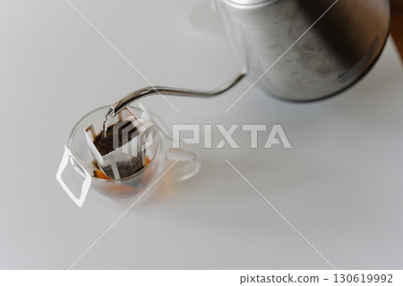 Close-up top view of pouring hot water from kettle into drip coffee bag nestled in glass cup, brewing fresh cup of coffee on white table. Process of making brewed coffee at home during breakfast. Close-up top view of pouring hot water from kettle into drip coffee bag nestled in glass cup, brewing fresh cup of coffee on white table. Process of making brewed coffee at home during breakfast. 130619992