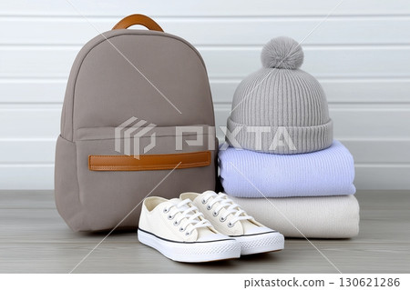 A neatly arranged stack of folded sweaters, a gray knitted hat with a pom-pom, a beige backpack 130621286