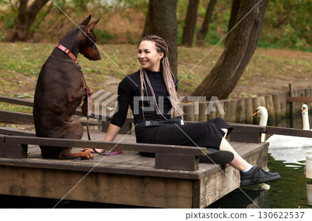 Woman sitting on dock with Doberman dog near scenic lake view Woman sitting on dock with Doberman dog near scenic lake view 130622537
