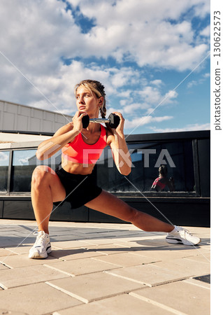 Young woman performing exercise with dumbbells outdoors under a bright sky Young woman performing exercise with dumbbells outdoors under a bright sky 130622573