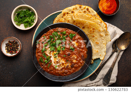 Traditional Indian Punjabi dish Dal makhani with lentils and beans in black bowl served with naan flat bread, fresh cilantro and two spoons on brown concrete rustic table top view 130622783