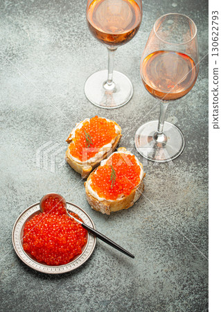 Small metal plate with red salmon caviar, two caviar toasts canape, two glasses of champagne top view on grey concrete background, festive luxury delicacy and appetizer Small metal plate with red salmon caviar, two caviar toasts canape, two glasses of champagne top view on grey concrete background, festive luxury delicacy and appetizer 130622793