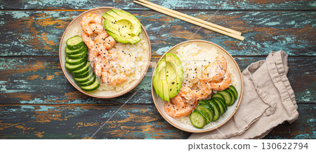 Two white ceramic bowls with rice, shrimps, avocado, vegetables and sesame seeds and chopsticks on colourful rustic wooden background top view. Healthy asian style poke bowl 130622794