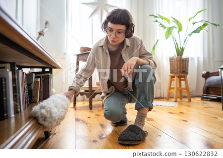 Tidy neat young woman using duster to remove dust from furniture near floor, home cleaning routine 130622832