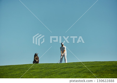 Golfer standing on hilltop with golf bag and club under blue sky 130622888