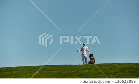Golfer walking across green hill carrying golf bag under bright sky 130622889