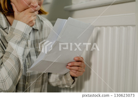 Upset woman holding utility paper bills next to heating thermostat, increasing heating costs 130623073