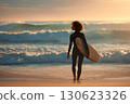 Beautiful young woman with surfboard standing on seashore with waves against sunset background 130623326