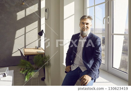 Mature businessman in suit sitting by the window in the office, looking at camera and smiling. Mature businessman in suit sitting by the window in the office, looking at camera and smiling. 130623419