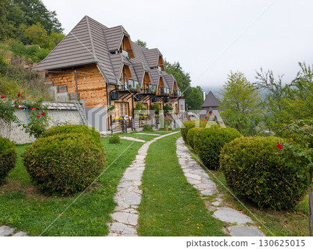 Row of small wooden cottages with pointed roofs, surrounded by trimmed bushes and stone path. Tourism and mountain accommodation in Serbia. 130625015