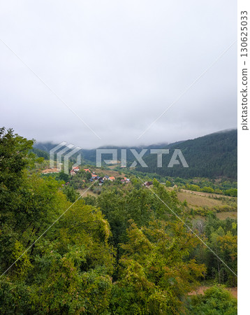 Foggy mountains and a valley with small houses and red roofs among green trees. Peaceful rural living and natural beauty in the Balkans. 130625033