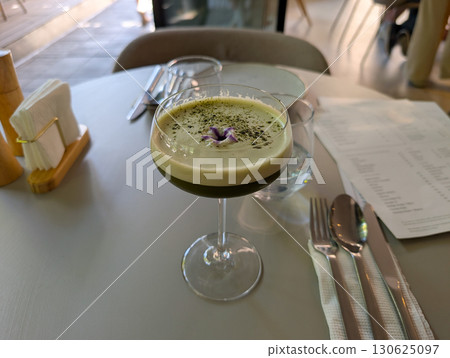 A green cocktail in a stemmed glass placed beside a menu on the table. Culinary presentation, lifestyle refreshment, and mixology art in dining culture. 130625097