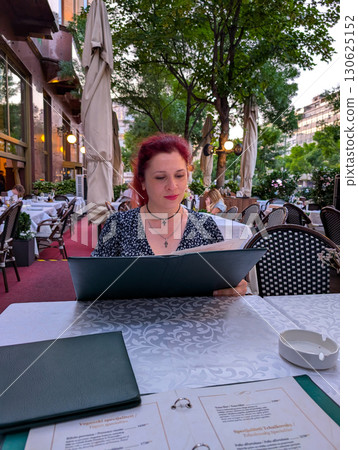 A woman reads a menu at an outdoor restaurant surrounded by tables and guests. Dining lifestyle, tourism, and evening leisure combining culture with culinary experience. 130625152