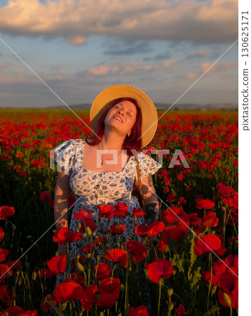 A woman smiles in a straw hat surrounded by red poppies at sunset. Lifestyle, beauty, and natural harmony of rural landscape with human presence and joy. 130625171