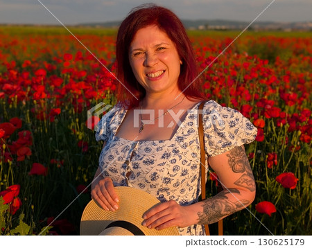 A woman smiles brightly in a poppy field with sunset light on her face. Portrait of happiness, individuality, and emotional lifestyle connection with summer nature. 130625179