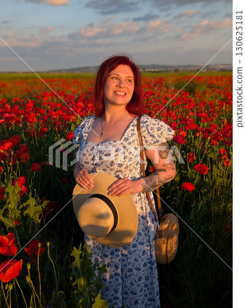 A woman smiles while holding straw hat in a poppy field at golden sunset. Lifestyle portrait, emotional charm, and the beauty of harmony between person and nature. 130625181