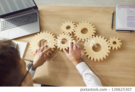 Top view of business man connecting big wooden gears together sitting at the desk on his workplace 130625307