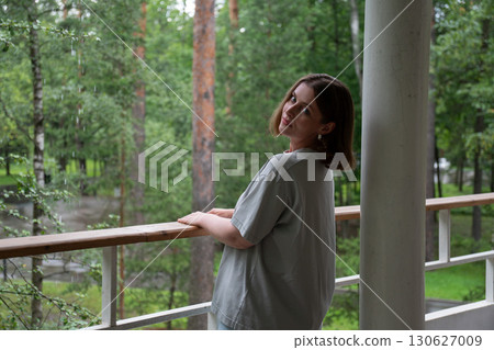 A woman in a casual grey t-shirt stands on a balcony in a green forest in Peredelkino, turning her head to look at the camera A woman in a casual grey t-shirt stands on a balcony in a green forest in Peredelkino, turning her head to look at the camera 130627009