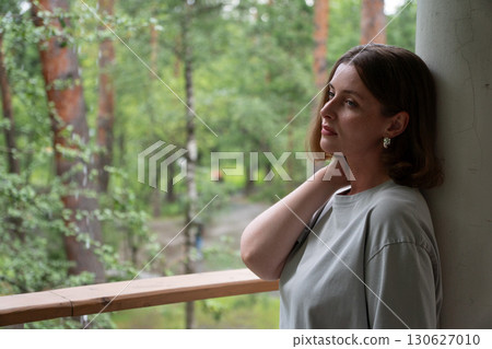 A woman leans against a column on a balcony in the forest in Peredelkino, gently holding her neck and gazing thoughtfully into the distance A woman leans against a column on a balcony in the forest in Peredelkino, gently holding her neck and gazing thoughtfully into the distance 130627010