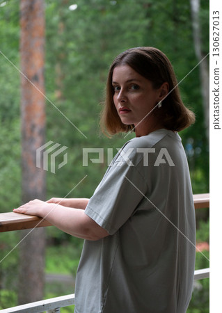 A serious young woman in a grey t-shirt stands on a balcony in a green forest in Peredelkino, looking thoughtfully into the camera A serious young woman in a grey t-shirt stands on a balcony in a green forest in Peredelkino, looking thoughtfully into the camera 130627013