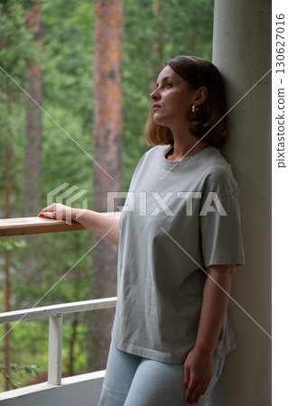 A young woman stands still on a forest balcony in Peredelkino, leaning against a column and looking up with a calm, distant gaze 130627016