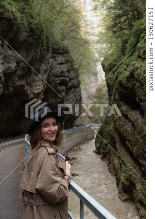 A young woman in a hat and trench coat smiles while walking along a canyon path with steep cliffs and a fast mountain river. solo travel, eco-tourism 130627151
