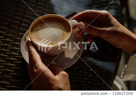 Coffee Hands Cup - Closeup of hands holding a cup of coffee with latte art on a patio table. Coffee Hands Cup - Closeup of hands holding a cup of coffee with latte art on a patio table. 130627208